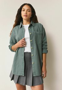 Groene denim shirt met knoopsluiting, kraag en borstzak, gecombineerd met een witte top en een grijze geplooide rok.