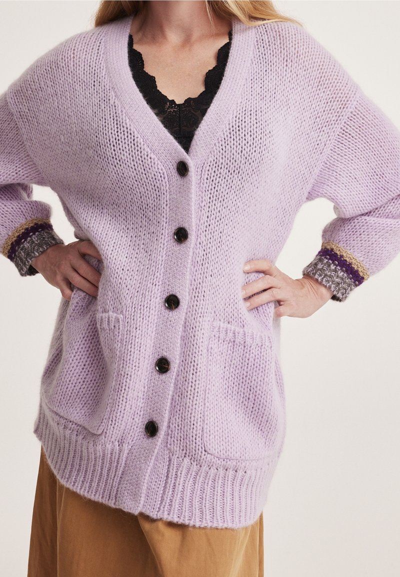 Femme portant un cardigan en maille lilas ample avec des boutons noirs, des poches plaquées et des poignets multicolores sur un haut en dentelle noire.