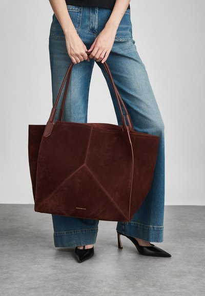 Καφέ σουέντ τσάντα tote με δομημένο σχέδιο, δερμάτινες λαβές και ανάγλυφο λογότυπο. Συνδυασμένη με ανοιχτό καμπάνα τζιν και μυτερές μαύρες γόβες.