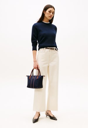 Marineblauer Pullover kombiniert mit cremefarbenen, weiten Hosen. Trägt eine marineblaue Handtasche mit braunen Griffen und pinken Streifen. Schwarze spitze Schuhe.