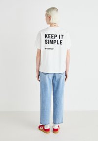 Hvit bomull T-skjorte med svart tekst "HOLD DET ENKELT MEN BETYDNINGSFULLT" på baksiden; paret med lyseblå jeans og røde sneakers.