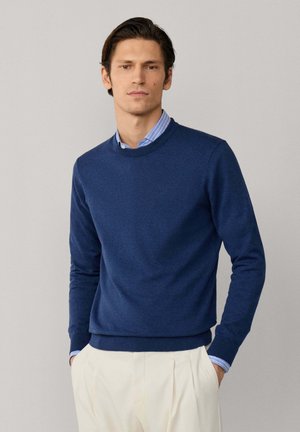 Man draagt een blauwe trui met ronde hals over een blauw gestreept overhemd met kraag, gecombineerd met crèmekleurige geplooide broek, staand tegen een effen grijze achtergrond.