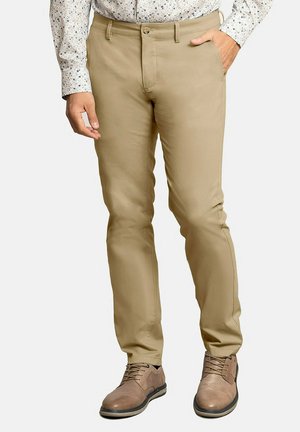 Chinos beige con un tejido suave y elástico, corte ajustado y bolsillos estándar, combinados con zapatos marrones y una camisa estampada.