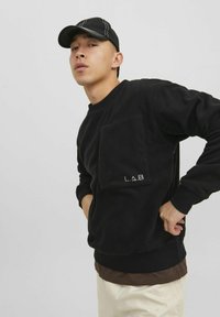 Čierna flísová mikina s veľkým predným vreckom a detailom loga. Model má na sebe čiernu čiapku, pričom predvádza jednoduchý, vrstvený outfit.