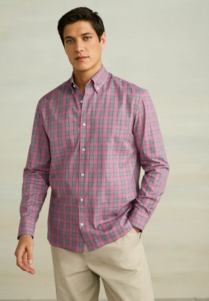 Mężczyzna w różowo-szarej, w kratę koszuli zapinanej na guziki oraz beżowych spodniach, stojący z jedną ręką w kieszeni na neutralnym tle.