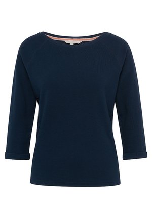 Kobieta ubrana w granatowy top, jasnoniebieskie obcisłe jeansy, srebrne trampki oraz małą niebieską torebkę na ramię, stoi z jedną nogą lekko ugiętą.