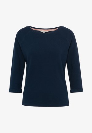 Kobieta ubrana w granatowy top, jasnoniebieskie obcisłe jeansy, srebrne trampki oraz małą niebieską torebkę na ramię, stoi z jedną nogą lekko ugiętą.
