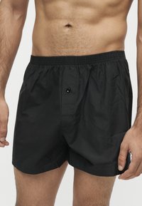 Torse masculin portant un boxer noir à taille élastique avec braguette à boutons, debout devant un fond clair uni.