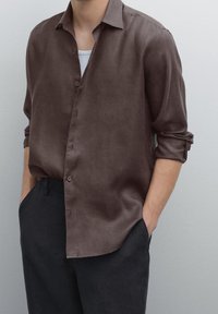Braunes Leinenhemd mit lockerem Schnitt, durchgehender Knopfleiste, hochgekrempelten Ärmeln und glatter Textur, kombiniert mit dunklen Hosen.