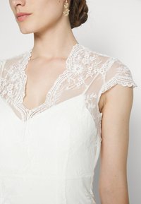Abito bianco con corpetto in pizzo dotato di un colletto a busto scallop e maniche a cappuccio, decorato con motivi floreali e tessuto testurizzato.