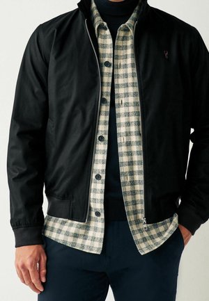 Homme portant une veste noire zippée sur une chemise à carreaux grise et blanche à boutons et un pull noir à col roulé.