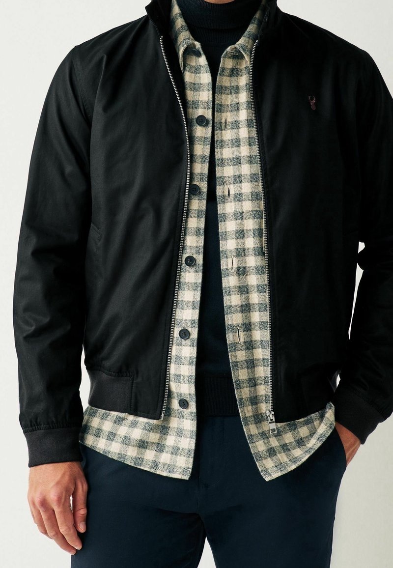Homme portant une veste noire zippée sur une chemise à carreaux grise et blanche à boutons et un pull noir à col roulé.