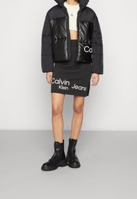 Giubbotto imbottito nero con pannelli lucidi, design accorciato, abbinato a una gonna nera con testo bianco "Calvin Klein Jeans" e stivali neri con zip.