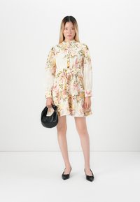 Donna in un vestito a fiori, con una borsa nera, che indossa tacchi a punta neri, in piedi davanti a uno sfondo bianco.