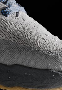 Šedá sneaker s texturovaným pleteným svrškem, kapky vody na povrchu, černá gumová podrážka a modré šněrování provlečené očky.