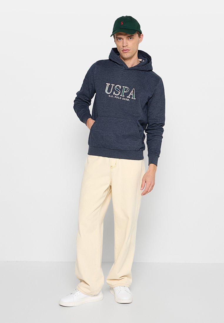 Homme portant un sweat à capuche bleu marine de USPA, un pantalon large crème, des baskets blanches et une casquette de baseball vert foncé, se tenant devant un fond clair.