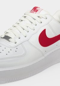 Λευκό αθλητικό παπούτσι Nike Air Force 1 με κόκκινο λογότυπο Swoosh, λευκά κορδόνια και κόκκινη ετικέτα "Nike Air Force 1" στη γλώσσα.