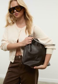 Store storebrune solbriller, beige cardigan med knapper og en mørk lædertaske med en sølvkædehåndtag.
