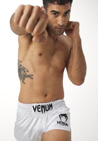 White satin shorts with black "Venum" logo and waistband, worn by a male model in a fighting stance, displaying tattoos and defined musculature.