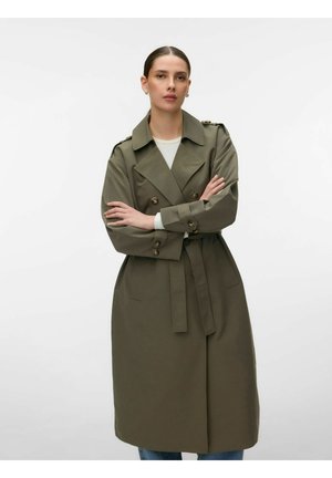 Femme aux cheveux plaqués en arrière portant un trench-coat vert olive croisé à double rangée de boutons avec une ceinture, debout les bras croisés devant un fond uni.