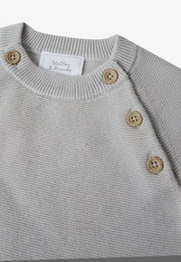Suéter de punto gris claro con un cuello acanalado, tres botones de madera en el hombro y una etiqueta tejida, creando una apariencia texturizada.