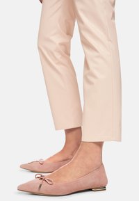 Rosa Wildleder-Ballerinas mit spitz zulaufender Zehenpartie, verziert mit einer Schleife und einem kleinen metallischen Akzent an der Ferse. Kombiniert mit hellen Hosen.