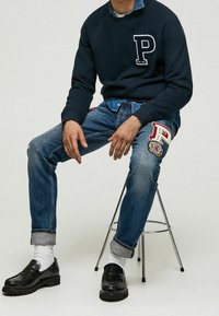 Sweatshirt marine avec logo "P", jeans en denim bleu avec des patchs sur les côtés, mocassins noirs et chaussettes blanches jusqu'aux chevilles, assis sur un tabouret en métal.