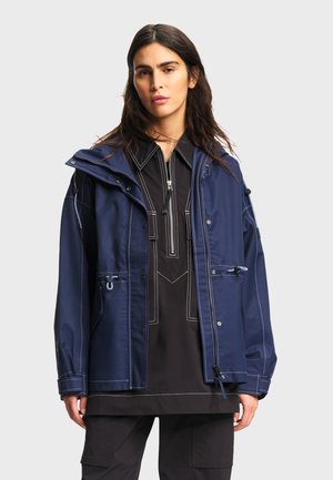 Woman with long dark hair wearing a navy blue jacket over a black zippered top and black pants against a plain light background.