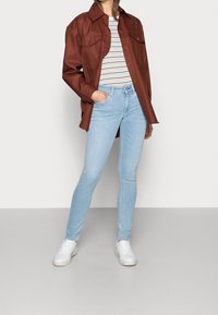 Persona con chaqueta marrón de botones, camisa a rayas, jeans ajustados azul claro, zapatillas blancas, de pie con una mano en el bolsillo.