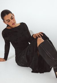 Melna garenroku kleita ar ziedu tekstūru, pieguļošs augšdaļa un plūstoša svārka daļa. Saderēta ar augstiem melniem ādas zābakiem. Sēž uz baltas virsmas.