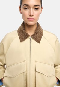 Kobieta w beżowej kurtce z dużymi przednimi kieszeniami i brązowym kołnierzem z kordurowej tkaniny, ręce w kieszeniach, neutralne tło.