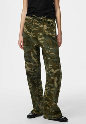 Person iført olivengrønne og brune camouflageprint løse bukser kombineret med sorte flade sko og en sort ærmeløs top.