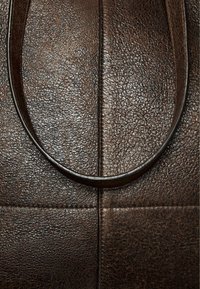 Bruine leren tas close-up, met een gestructureerd oppervlak, rechthoekige panels en een ronde handgreep. Het materiaal lijkt duurzaam en zacht.