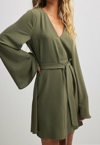 Frau in olivgrünem, tailliertem Kleid mit breiten Glockenärmeln und V-Ausschnitt, Hand auf der Hüfte, vor hellem, einfarbigem Hintergrund.
