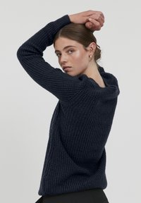 Marineblauer Strickpullover mit strukturiertem Finish, der einen weiten Kragen und lange Ärmel aufweist, von hinten gezeigt, während das Arm des Models erhoben ist.