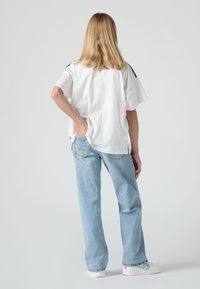 Osoba s plavom kosom stoji okrenuta leđima, obučena u bijelu majicu, svijetloplave traperice, bijele tenisice, s jednom rukom u stražnjem džepu.