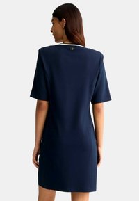 Vestito blu navy a maniche corte con taglio dritto, caratterizzato da un colletto bianco e un piccolo logo sulla parte posteriore. Tessuto dalla texture liscia.