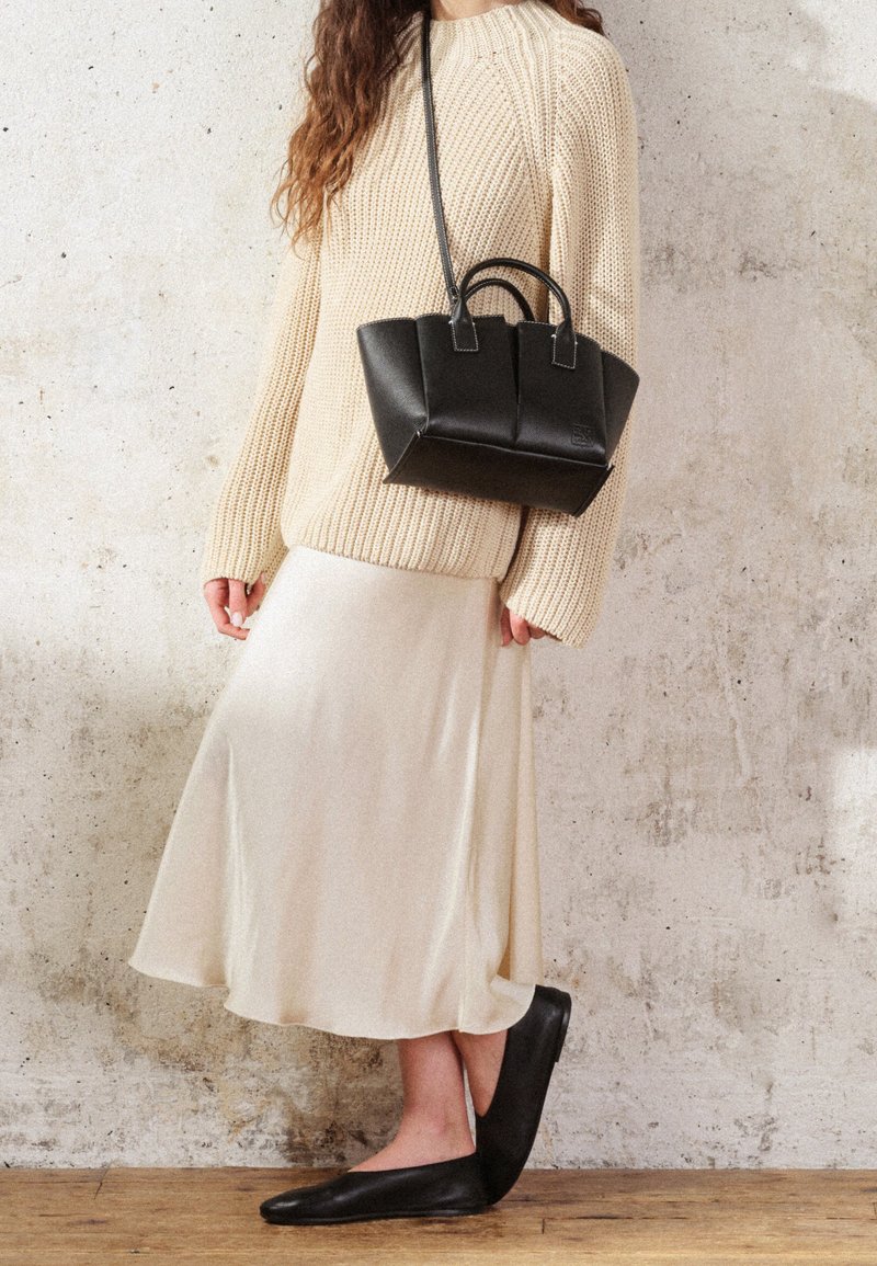 Borsa in pelle nera con un design strutturato, abbinata a un maglione lavorato a maglia crema e una gonna fluida crema. Scarpe basse nere sotto.