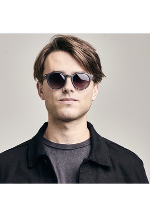 Ronde zonnebrillen met donkere lenzen en een grijze omlijsting, gedragen door een persoon in een zwarte jas over een grijze shirt. Neutrale achtergrond.