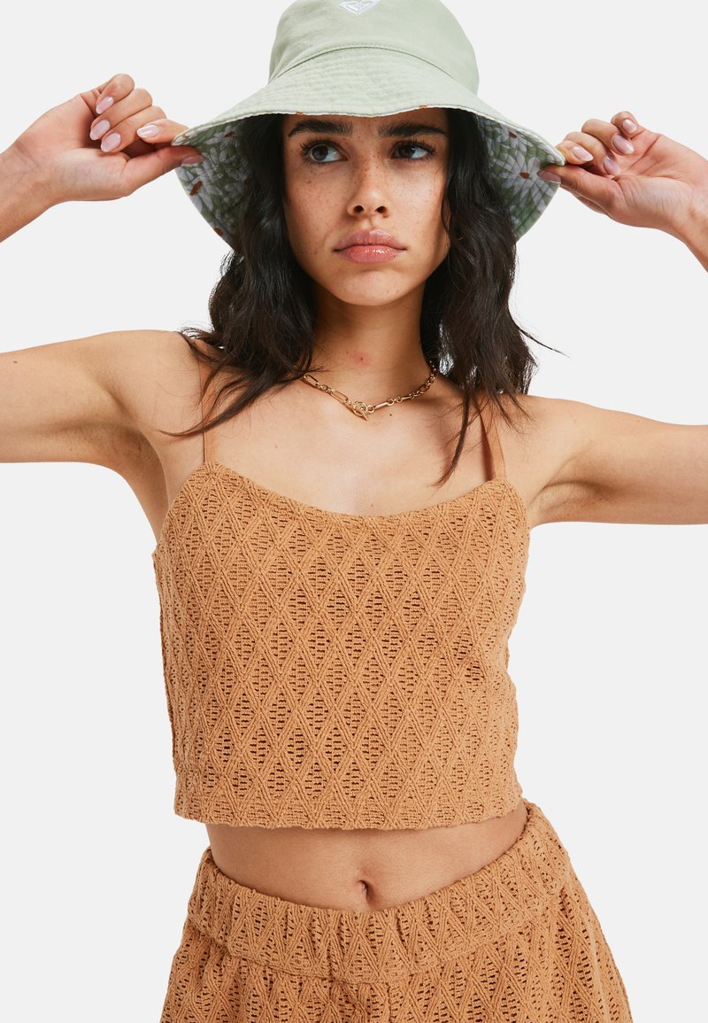 Woman adjusting a light green bucket hat, wearing a tan crochet patterned crop top and matching shorts, with a gold chain necklace.