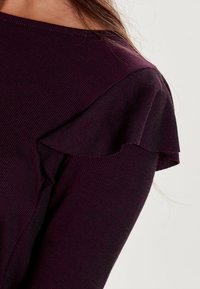 Dlouhý rukáv top v tmavě fialové proužkované tkanině. Obsahuje volánkový detail na rameni a široký výstřih. Hladká textura a přiléhavý design.