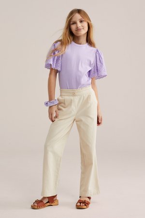 Niña joven con una blusa lavanda de mangas abullonadas, pantalones anchos color crema, sandalias marrones y una goma de pelo lavanda a juego en la muñeca.