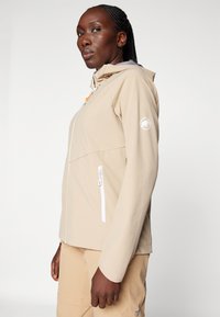 Beige softshelljacka med huva, utrustad med dragkedja fram, vita dragkedjefickor och en logotyp på ärmen. Slät textur.