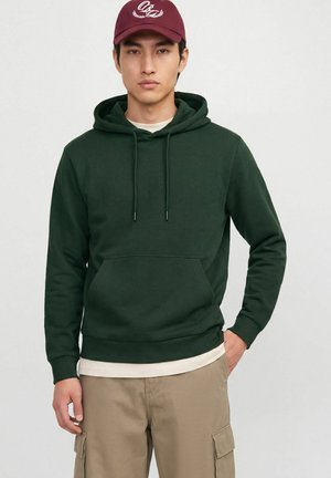 Jeune homme portant une casquette bordeaux, un sweat à capuche vert foncé avec une poche avant, un pantalon cargo beige, debout devant un fond gris clair uni.