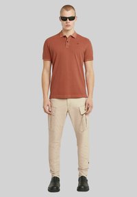 Polo shirt color ruggine con una chiusura a tre bottoni, abbinato a pantaloni cargo beige con multiple tasche e una vestibilità slim. Stivali neri.