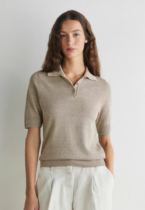 Kvinde iført en beige strikket polo med korte ærmer, stukket ned i højtaljede hvide bukser, stående foran en almindelig lys væg.