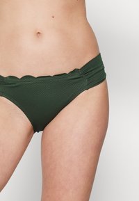 Parte de baixo de bikini verde escuro com um padrão texturizado, bordas onduladas e detalhes franzidos nas laterais. Composta por um material de banho elástico.