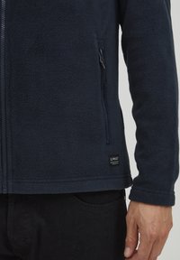 Chaqueta de forro polar azul marino con un bolsillo con cremallera, tela texturizada y una etiqueta de logo en el dobladillo. Además, cuenta con un cuello alto y puños ajustados.