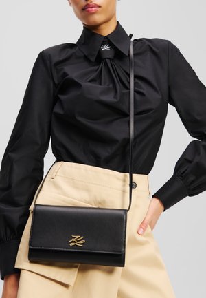 Sac bandoulière en cuir noir avec un logo doré, de forme rectangulaire, texture lisse et une longue sangle, porté avec une jupe beige et un chemisier noir.