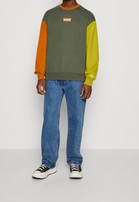 Felpa a blocchi di colore in verde oliva, arancione e verde lime con logo Levi's. Abbinata a jeans di denim blu e scarpe da ginnastica nere.